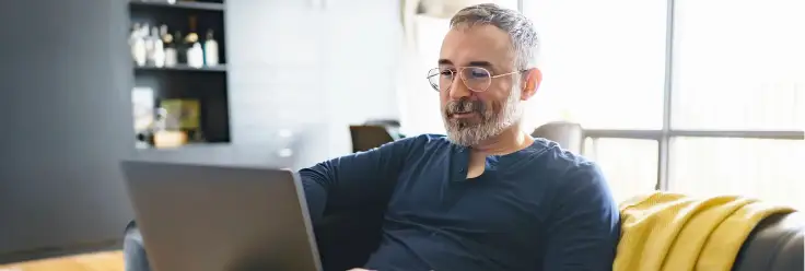 Middle-aged man sitting on a couch looking at a laptop screen.